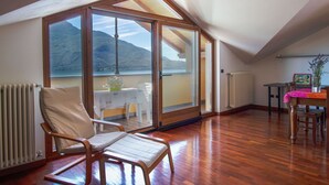 A spacious room with a wooden floor, a large sliding glass door leading to a balcony, a chair, a table, and a radiator. - Cosy apartment for 4 guests with WIFI, TV and terrace (Domaso)