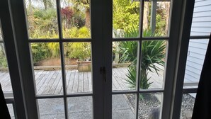 Une vue à travers une fenêtre donnant sur une terrasse en bois, entourée de différentes plantes et bordée d’un chemin en pierre.