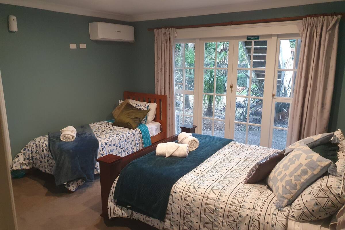 A bedroom with two beds, a window with curtains, an air conditioning unit, and a view of greenery outside.