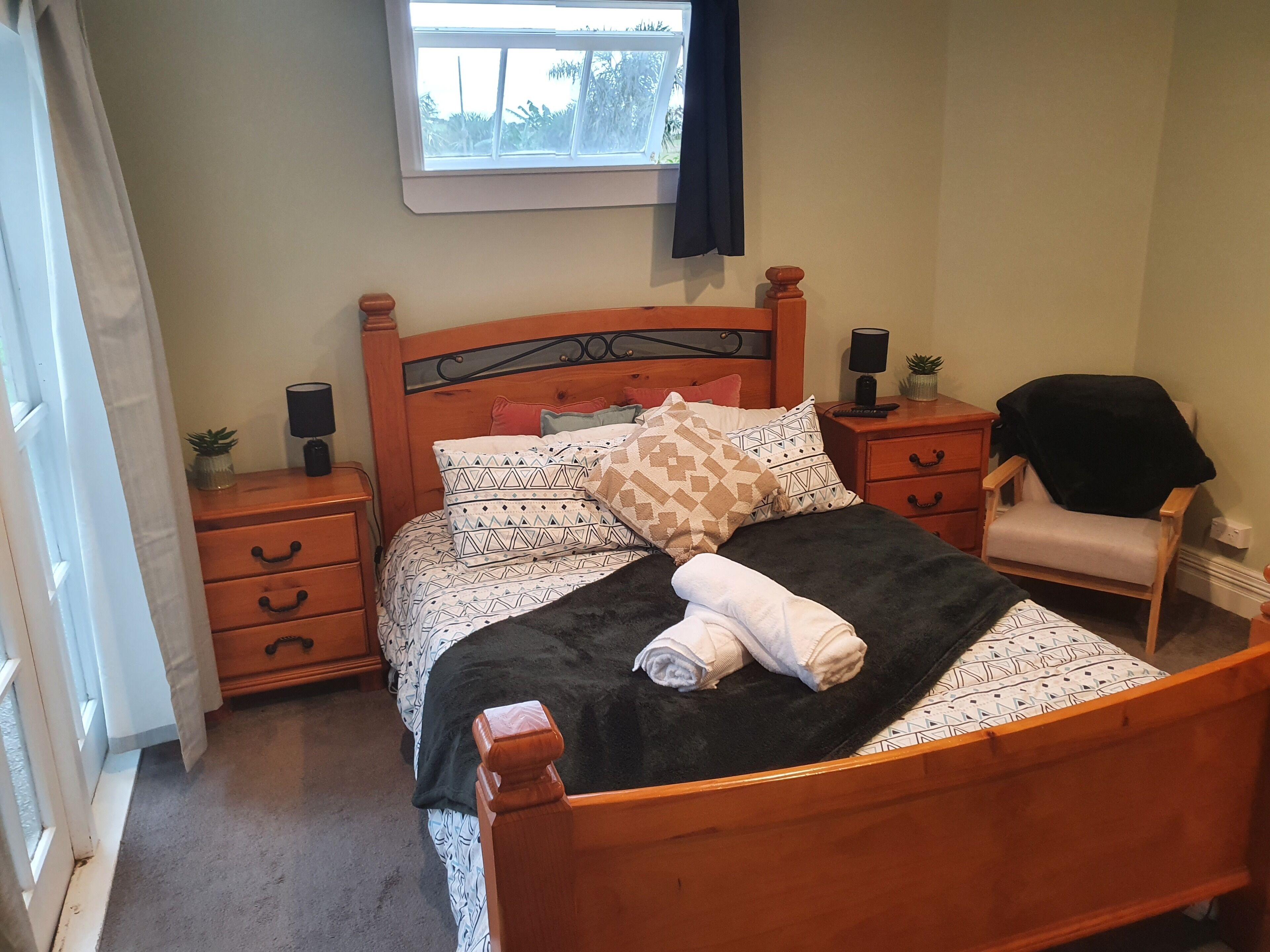 A neatly made bed with patterned bedding, a bedside table with a lamp, a chair, and a window with curtains.