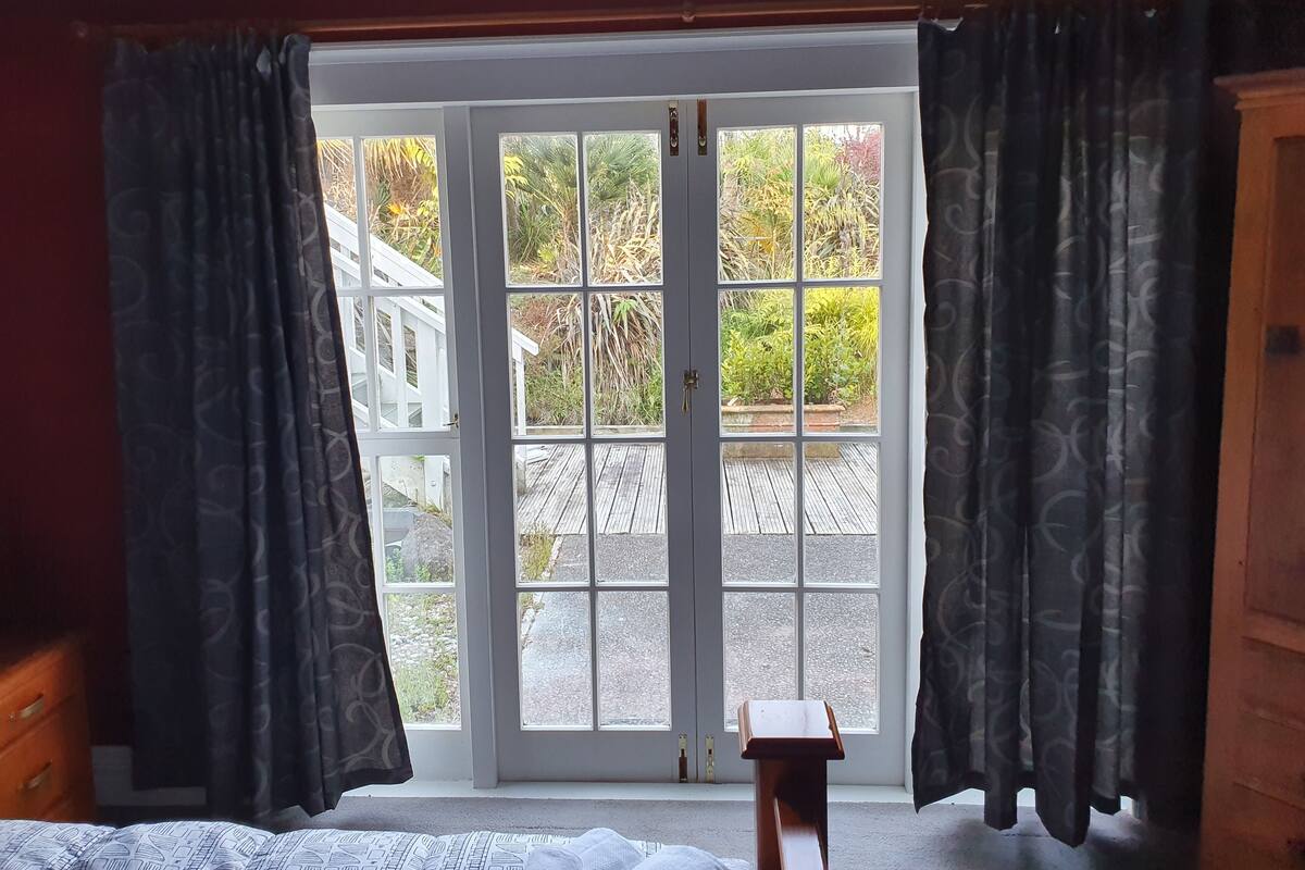 A room with a bed, a wooden desk, and a large sliding glass door leading to an outdoor area with a wooden deck and greenery.