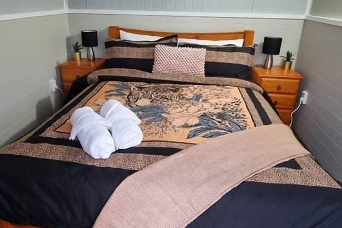 A neatly made bed with a patterned bedspread, two towels, and a blanket. There are two bedside tables with lamps and a small plant on each. The room has a simple, clean design with a grey carpet.
