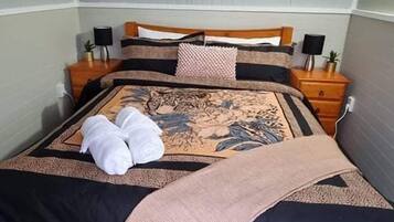 A neatly made bed with a patterned bedspread, two towels, and a blanket. There are two bedside tables with lamps and a small plant on each. The room has a simple, clean design with a grey carpet.
