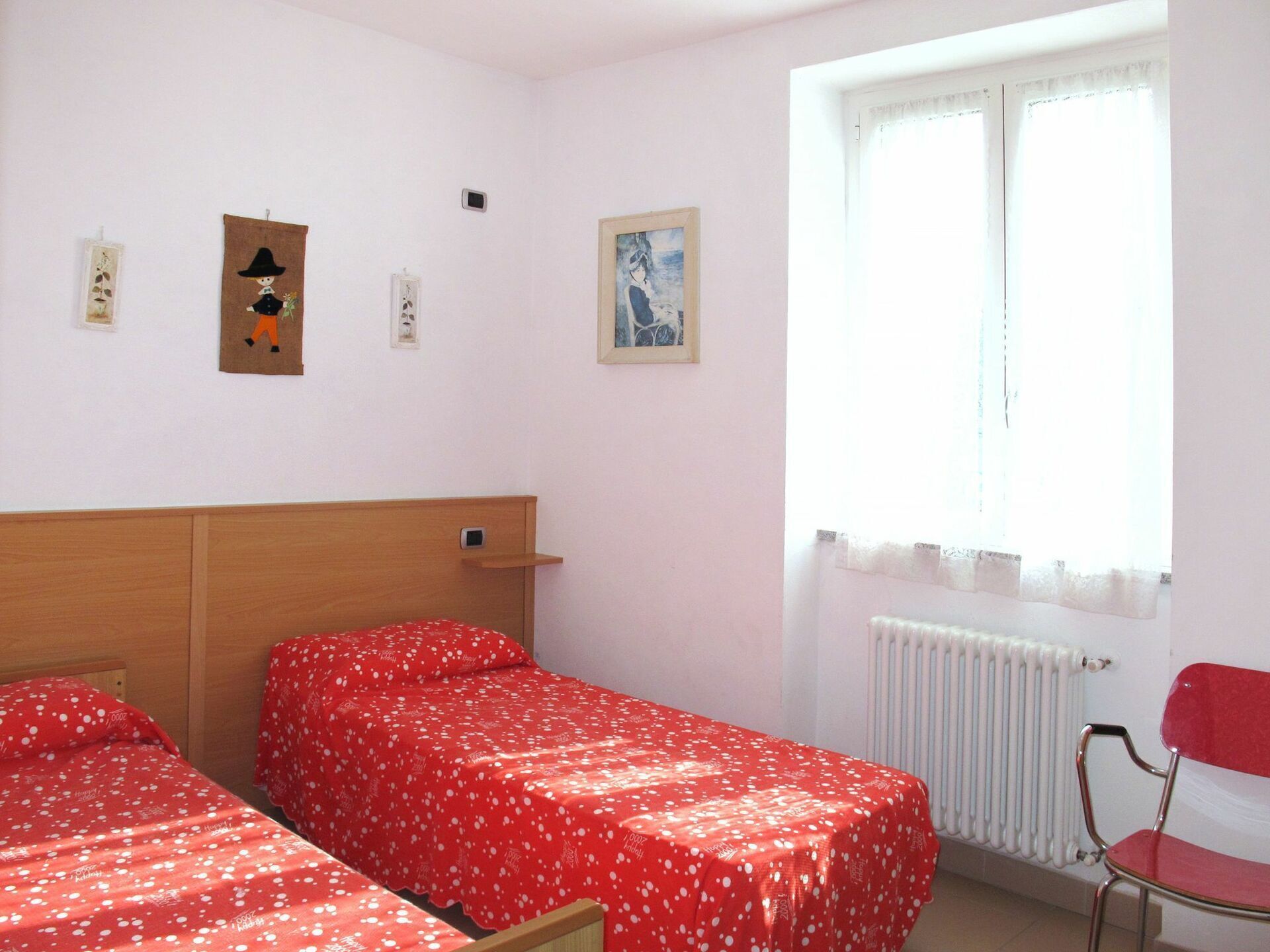 Une chambre avec deux lits simples, une tête de lit en bois, un radiateur, une fenêtre avec des rideaux et une chaise rouge.