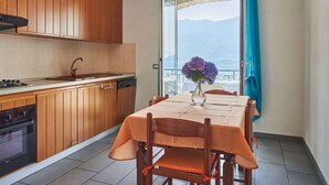 A kitchen with wooden cabinets, a dining table with chairs, a vase with flowers, and a view of mountains through a window. - Cosy apartment for 4 people with WIFI, TV, terrace and pets allowed (Vercana)
