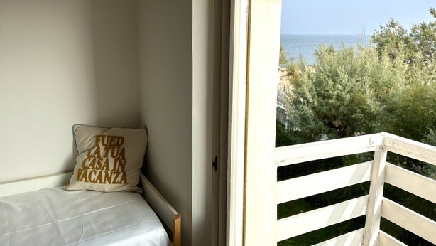 A balcony with a white railing, a bed with a white mattress and a pillow with text, and a view of the sea and greenery. - M150 - Marcelli, two-room apartment directly on the beach (Marcelli)