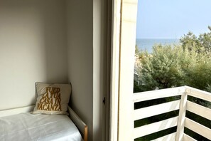 Een balkon met een witte balustrade, een bed met een witte matras en een kussen met tekst, en uitzicht op zee en groen.