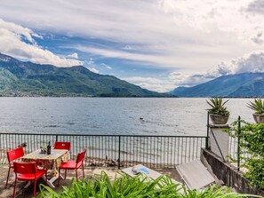 A lakeside terrace with red chairs and a table, overlooking a lake with mountains in the background. - Beautiful apartment for 4 guests with WIFI, TV, terrace and pets allowed (Gera Lario)