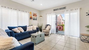 A living room with a blue sofa, a small table, and a view of a pink house through a glass door. - 5-Min Walk to Beach | Pool | Loaded Game Room | Near Shops & Outlets ✨ (Miramar Beach)