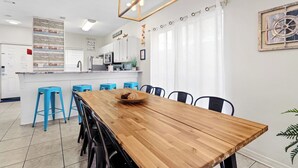 A modern kitchen with white cabinets, a wooden dining table, and blue bar stools. - 5-Min Walk to Beach | Pool | Loaded Game Room | Near Shops & Outlets ✨ (Miramar Beach)