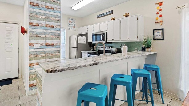 A modern kitchen with white cabinets, a marble countertop, and blue bar stools. - 5-Min Walk to Beach | Pool | Loaded Game Room | Near Shops & Outlets ✨ (Miramar Beach)