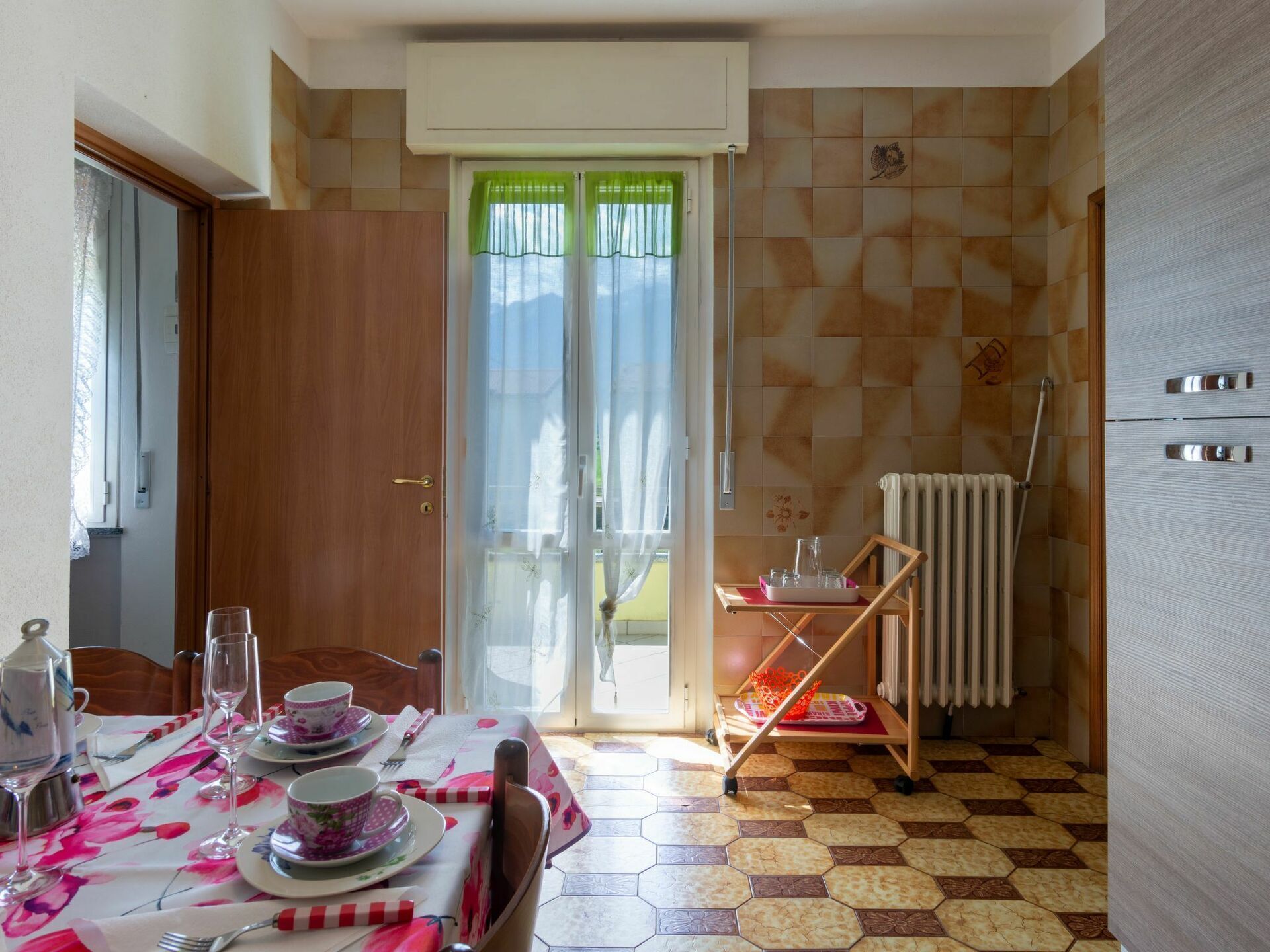 Uma sala de jantar com uma mesa posta para quatro pessoas, um radiador e uma porta com cortinas verdes.