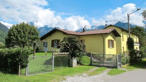 Ein gelbes Haus mit rotem Dach, umgeben von Grünflächen und Bergen im Hintergrund.