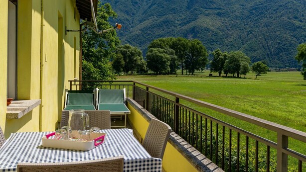 Ein Balkon mit kariertem Tischleinen, Korbstühlen und einem Tisch, gedeckt mit einer Kanne und Gläsern. Im Hintergrund ist ein bewaldeter Berg in der Ferne sichtbar.
