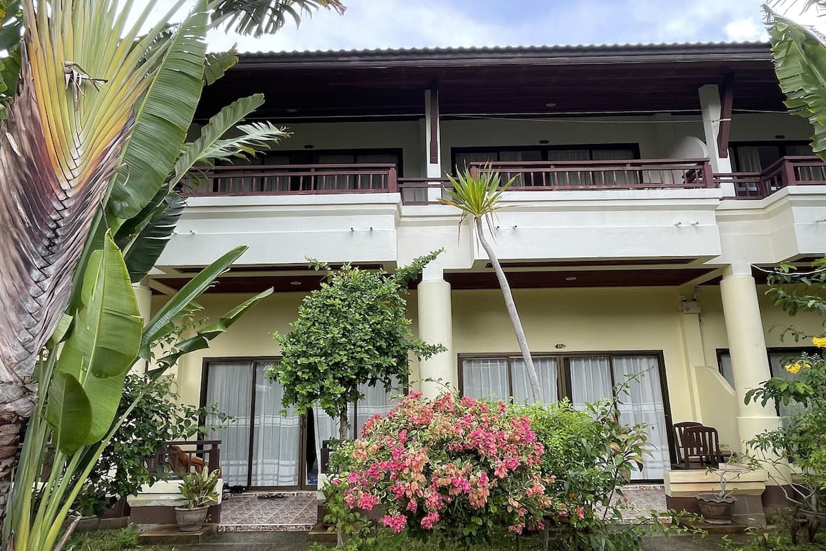 A two-story building with a balcony, surrounded by tropical plants and a small garden.
