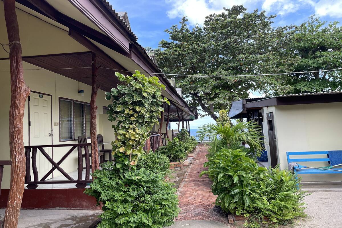 A covered walkway with a herringbone tile pattern, flanked by lush greenery and a blue bench.