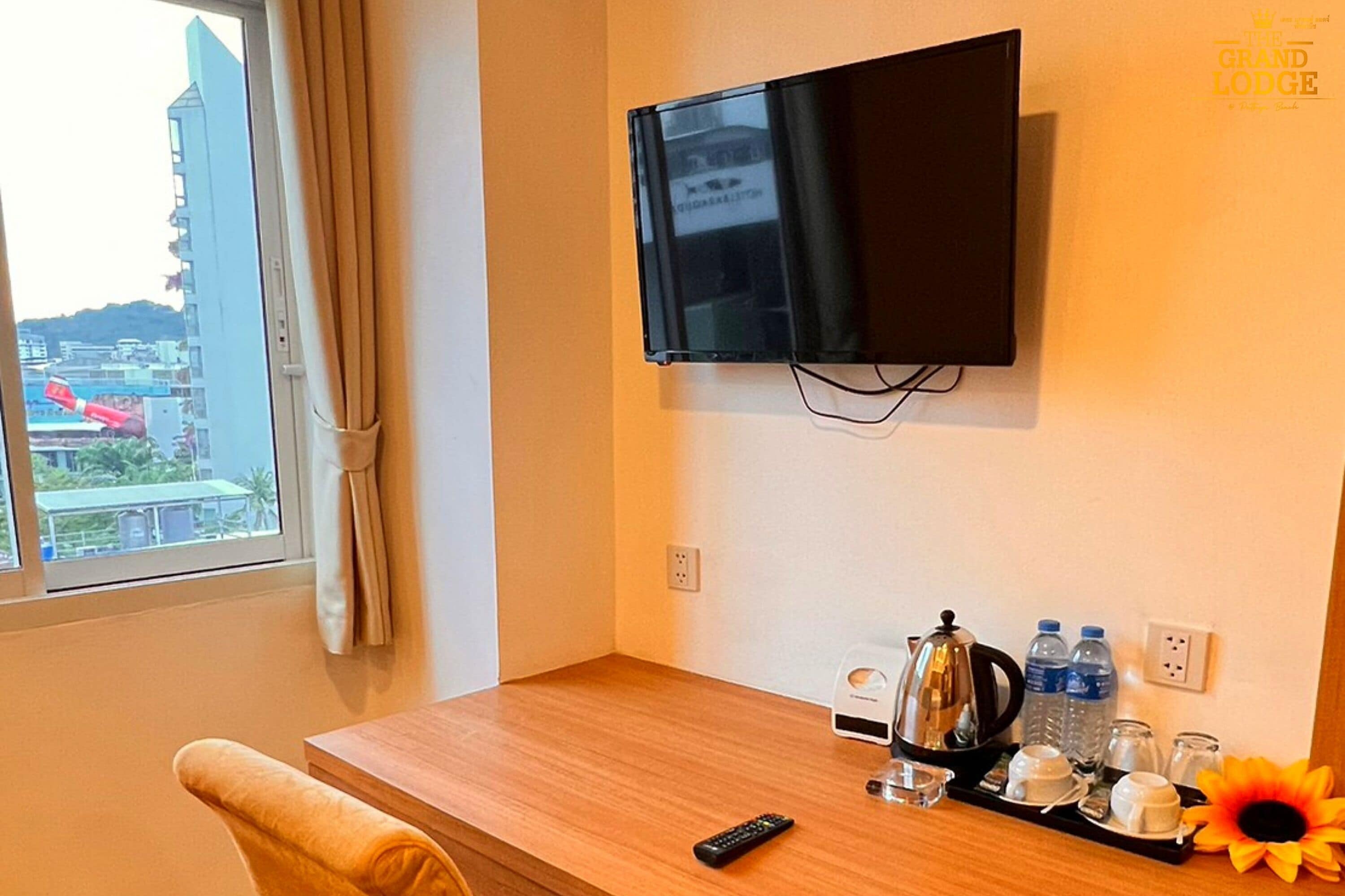 Ein Hotelzimmer mit an der Wand montiertem Fernseher, einem Holztisch mit Fernbedienung, einem Tablett mit Tassen und einem Wasserkocher sowie einem Fenster mit Blick auf Gebäude und eine rote Struktur im Freien.