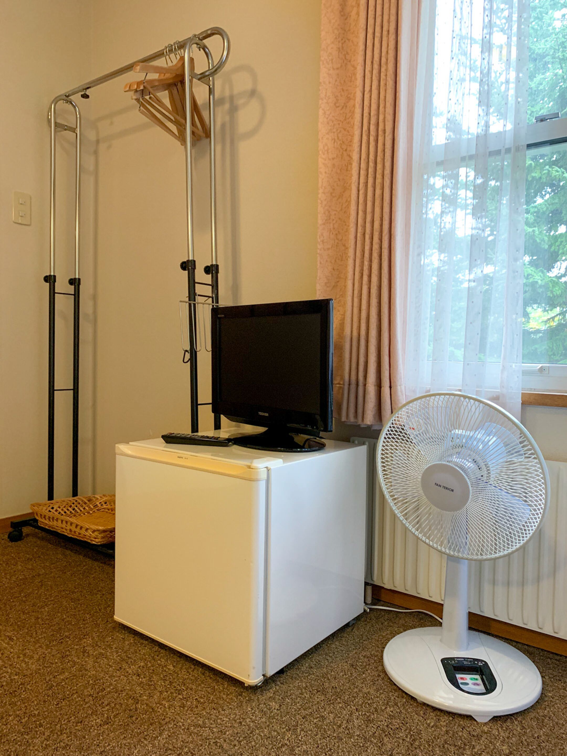 A room with a white washing machine, a black monitor on top, a standing fan, and a window with curtains.