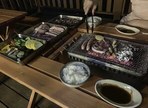 A barbecue setup with various meats and vegetables on a grill, a bowl of rice, and a plate of dipping sauce.