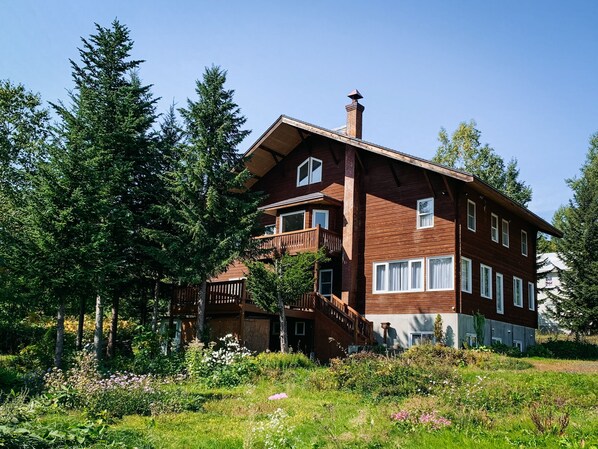 A two-story wooden house with a balcony, surrounded by trees and a garden.