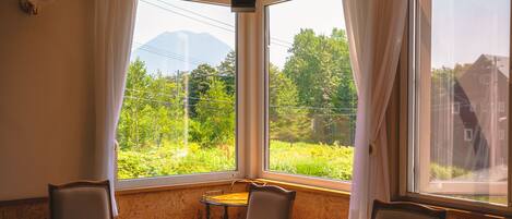 A dining area with a table and chairs, large windows with curtains, and a view of greenery and buildings.