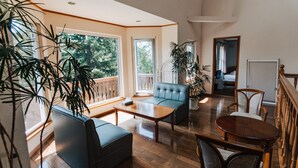 A spacious living area with a wooden floor, a blue sofa, a wooden coffee table, and a dining table with chairs. There are large windows with a view of trees and a balcony. - The Niseko Ski Lodge - Higashiyama (Niseko)