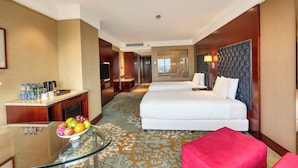 Une chambre d’hôtel avec un lit, un bureau sur lequel se trouve un panier de fruits, une chaise rouge et une vue sur le couloir.