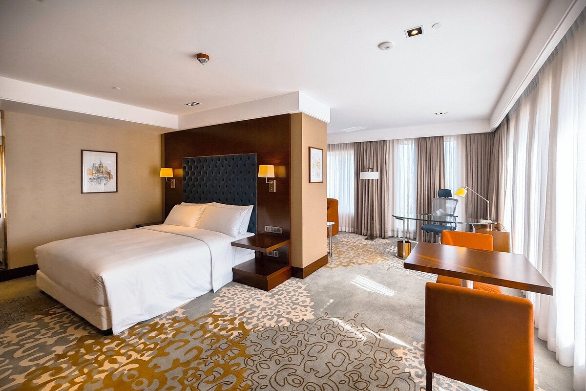 A hotel room with a large bed, a desk, a chair, and a view of the city through the window.