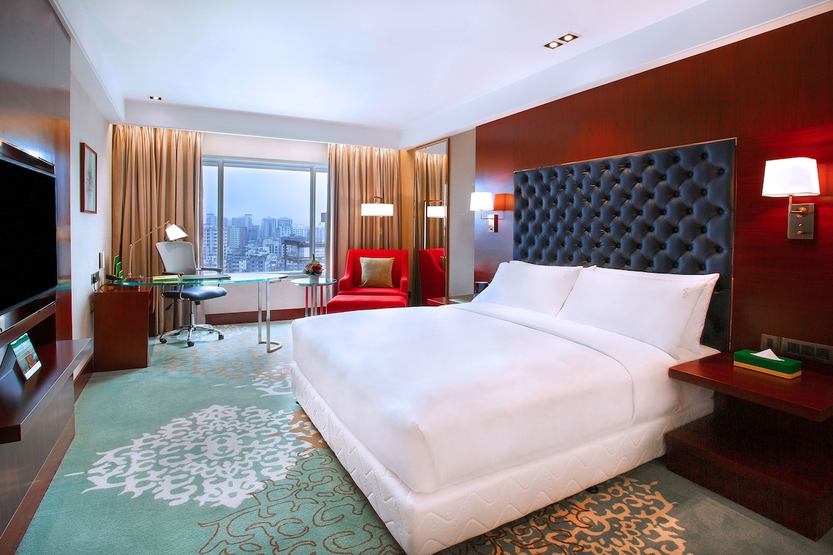 A hotel room with a large bed, a desk, a red chair, a television, and a city view.