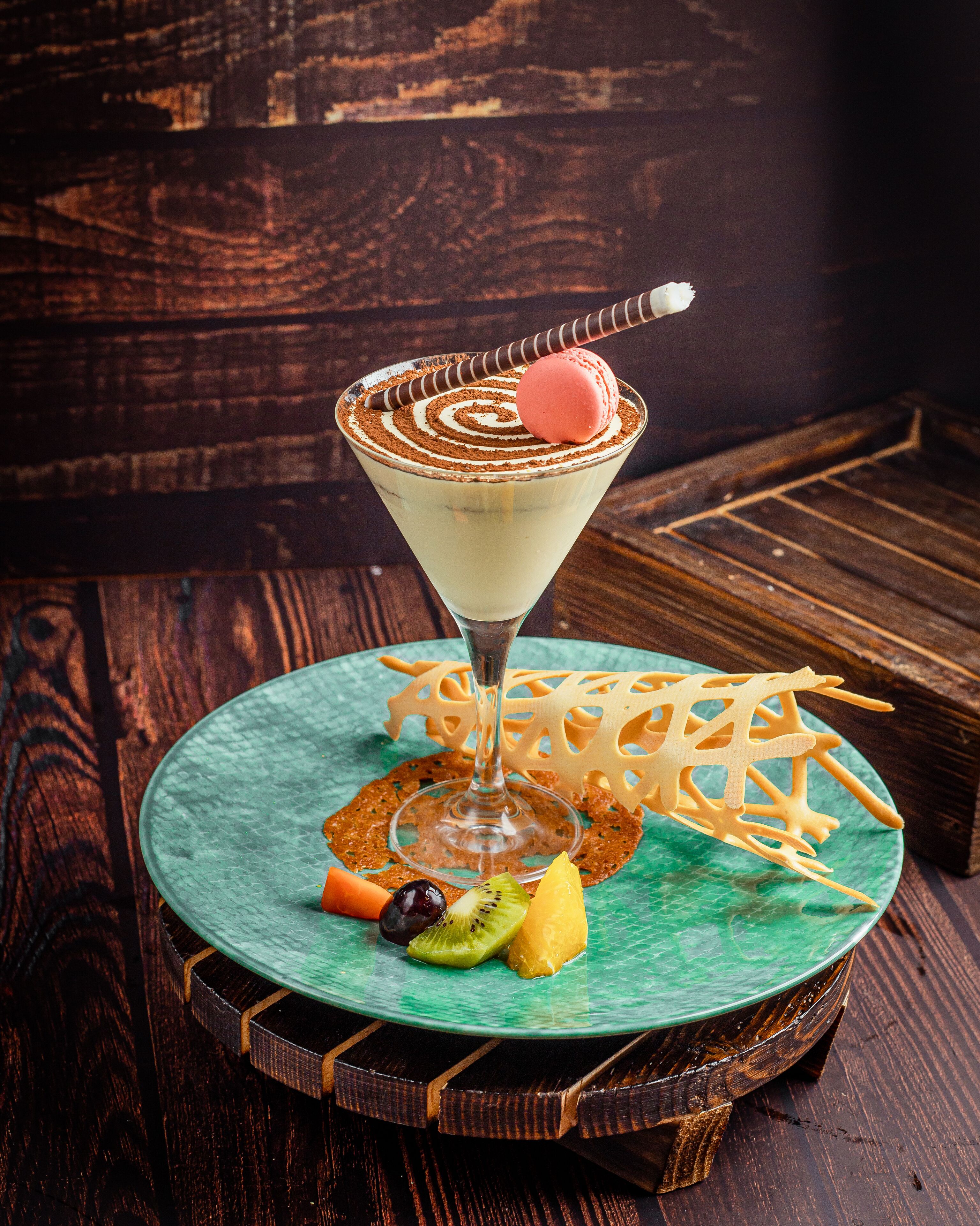 Una copa de martini con un remolino de chocolate y un macaron rosa sobre un plato verde, rodeado de fruta y un adorno de madera sobre una mesa de madera.