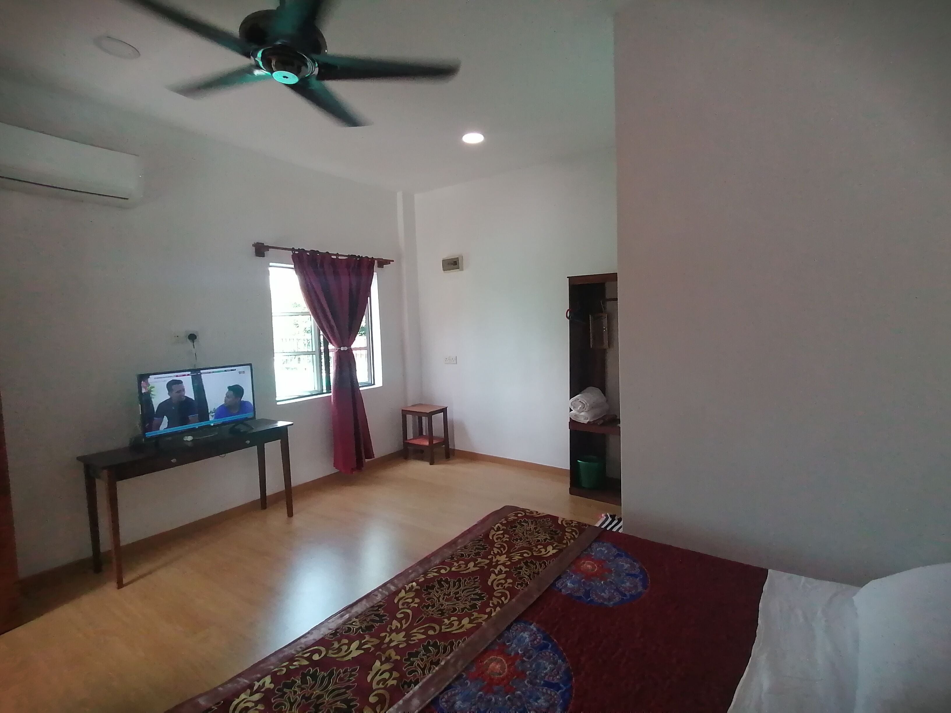 A living room with a ceiling fan, a television on a stand, a window with curtains, a small wooden table, a patterned rug, and a green trash bin.