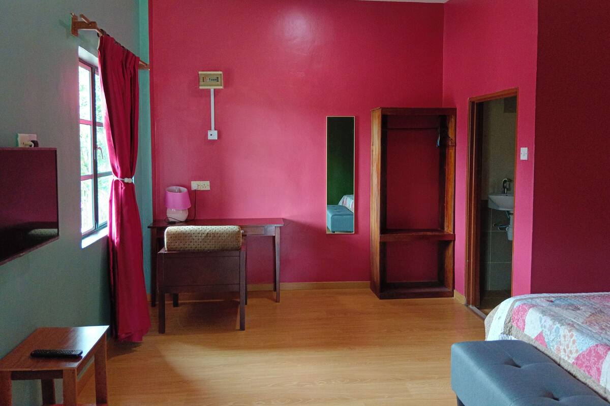 A room with a bed, a desk, and a chair. There is a window with curtains, a mirror, and a door leading to a bathroom.