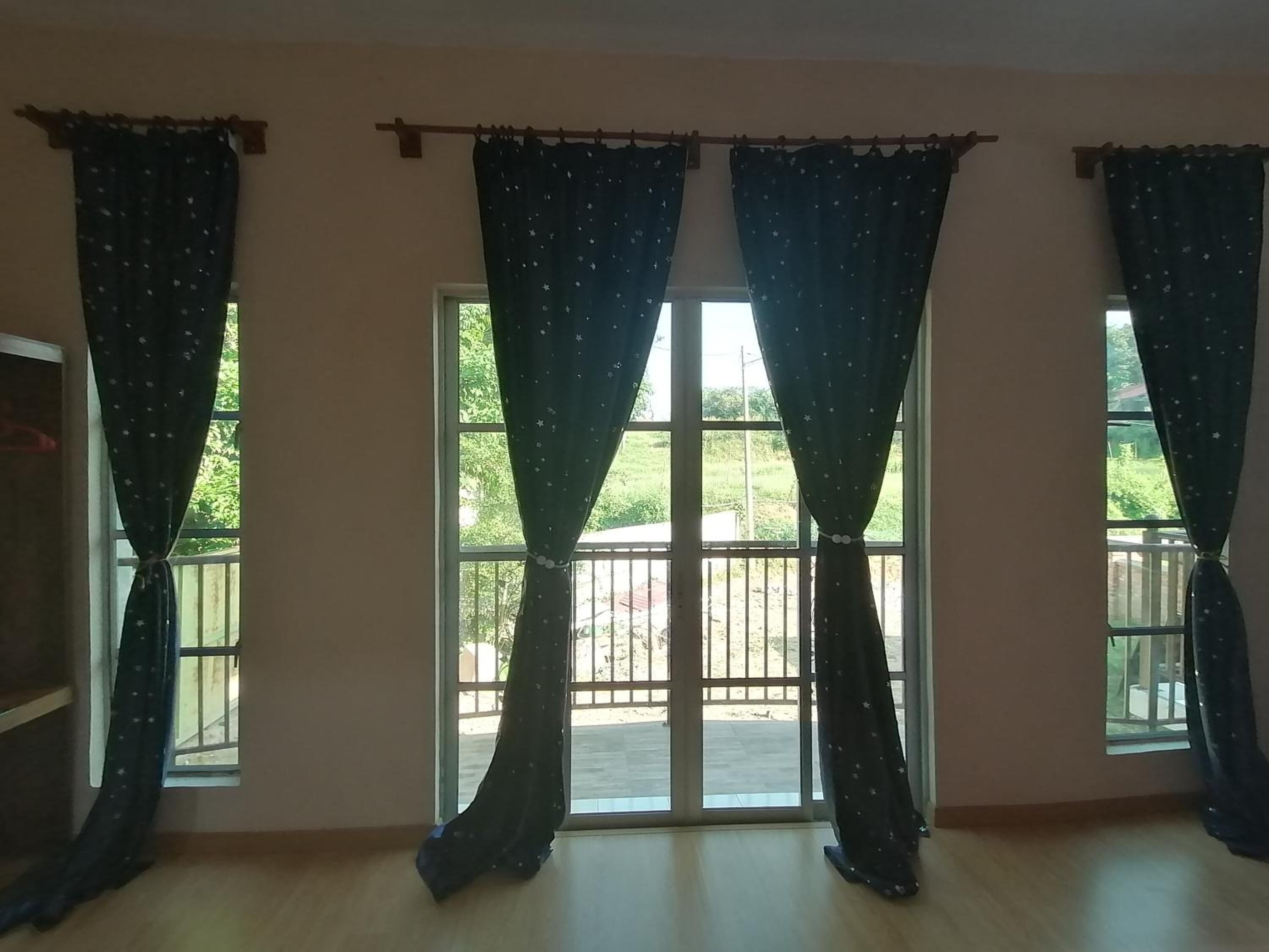 A room with three windows, each with dark curtains featuring a star pattern. The windows are large and open, revealing a view of a balcony with a railing.