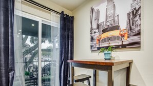 A modern room with a sliding glass door leading to a balcony, a wooden table with two stools, and a cityscape painting on the wall. - Stylish Condo near SMU, NorthPark & Greenville Ave with Pool (Dallas)