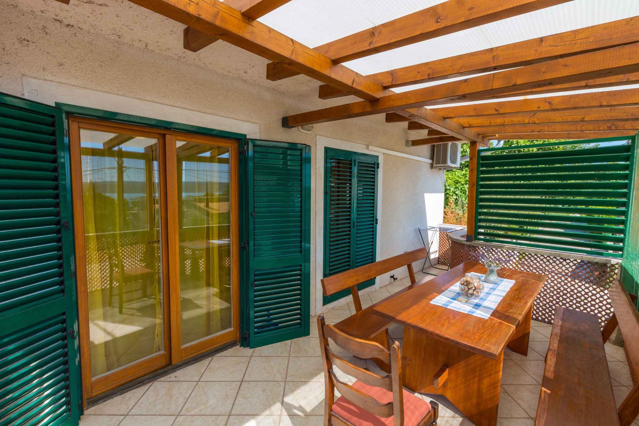 Eine überdachte Terrasse mit einem Holztisch und -stühlen, grünen Rollläden und Blick ins Grüne.