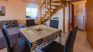Un coin repas avec une table en verre, quatre chaises, un canapé agrémenté d’un coussin à motifs floraux, un escalier en bois et une cuisine équipée d’un réfrigérateur.