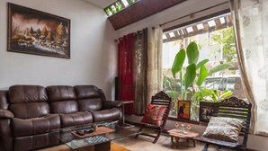 A living room with a brown leather sofa, a glass coffee table, and a wooden side table. There is a large window with curtains, a plant, and a framed picture on the wall.