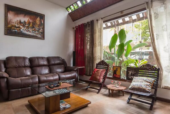 A living room with a brown leather sofa, a glass coffee table, and a wooden side table. There is a large window with curtains, a plant, and a framed picture on the wall.