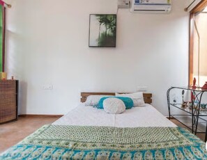 A bedroom with a bed, a wall-mounted air conditioner, a framed picture, and a metal shelf with decorative items.