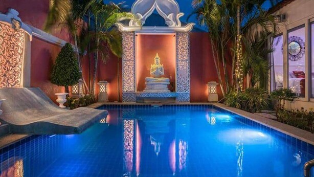 A swimming pool with a decorative structure in the center, surrounded by palm trees and a building with patterned walls. - View Talay Villas Pattaya (Pattaya)