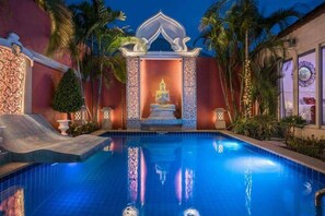 A swimming pool with a decorative structure in the center, surrounded by palm trees and a building with patterned walls. - View Talay Villas Pattaya (Pattaya)