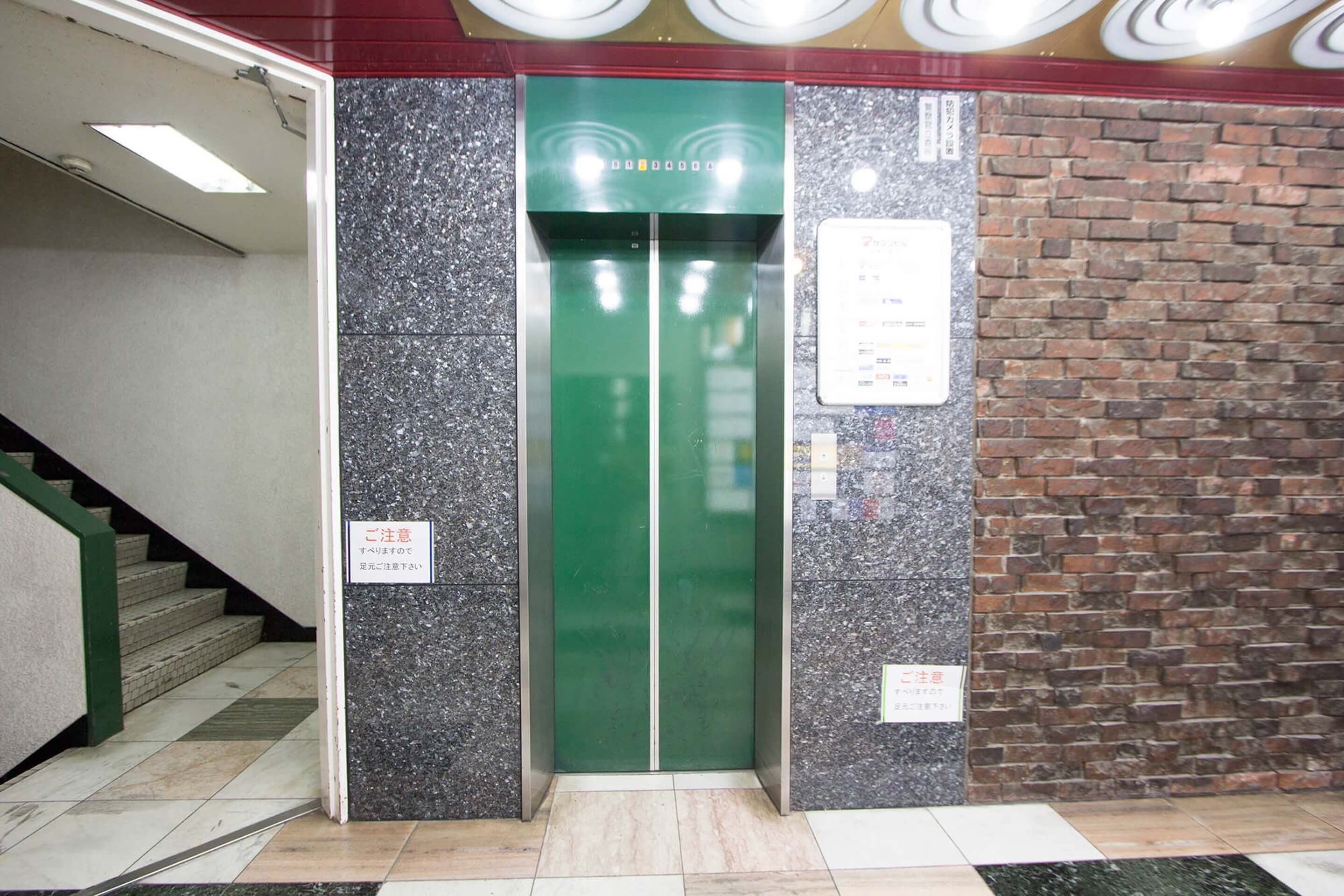 An elevator with a green door, a staircase to the left, and a brick wall to the right.