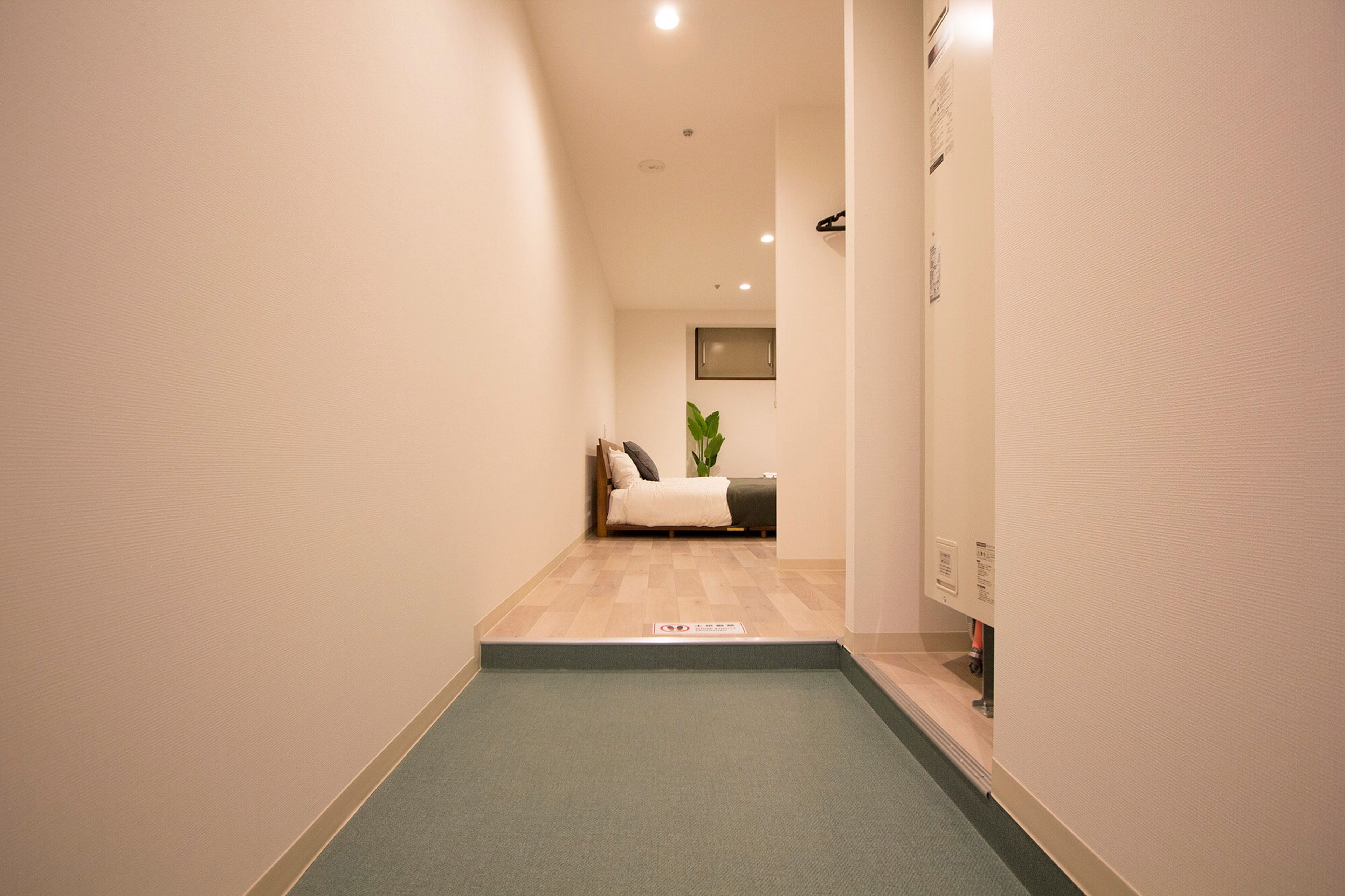 A narrow hallway with a green carpet leading to a room with a bed and a plant.