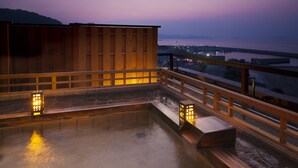Zona de baños termales con barandillas de madera y escalones de piedra, con vistas a una masa de agua y luces de la ciudad a lo lejos.