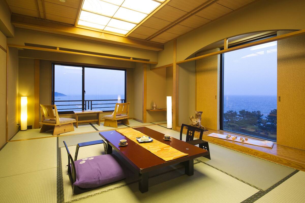 Une pièce de style japonais traditionnel, avec un sol recouvert de tatamis, une table basse, des chaises et une grande fenêtre donnant sur la mer.