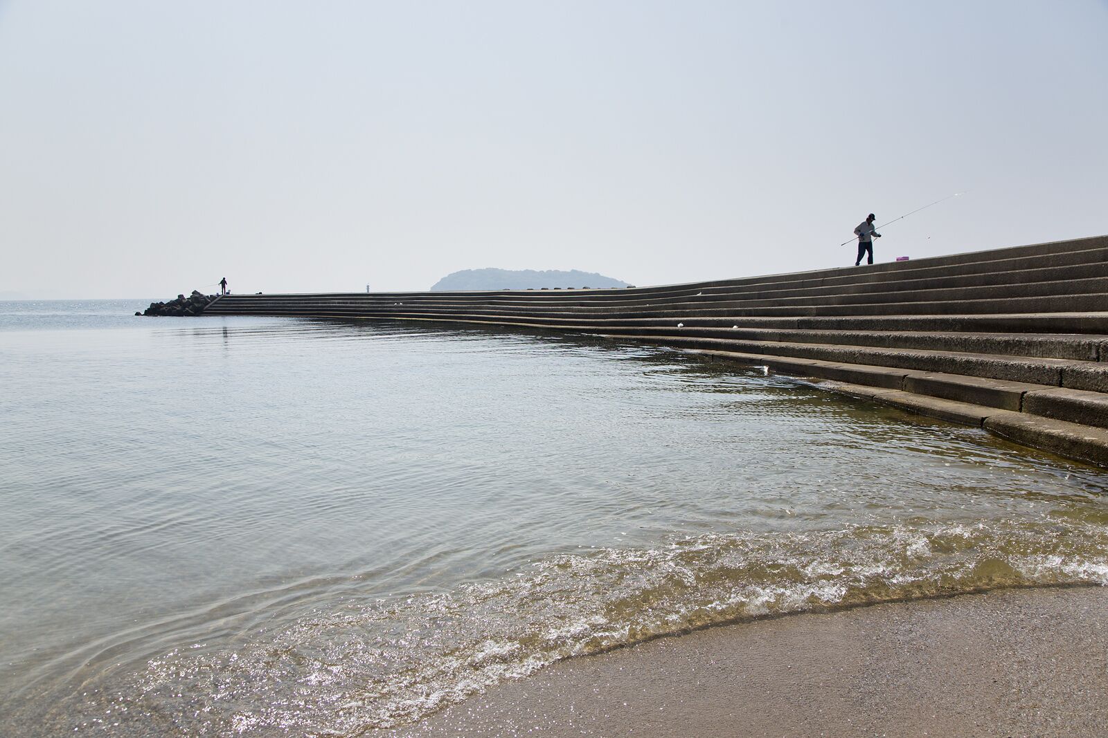 A waterfront promenade with steps leading into the water, a person fishing, and a distant mountain.