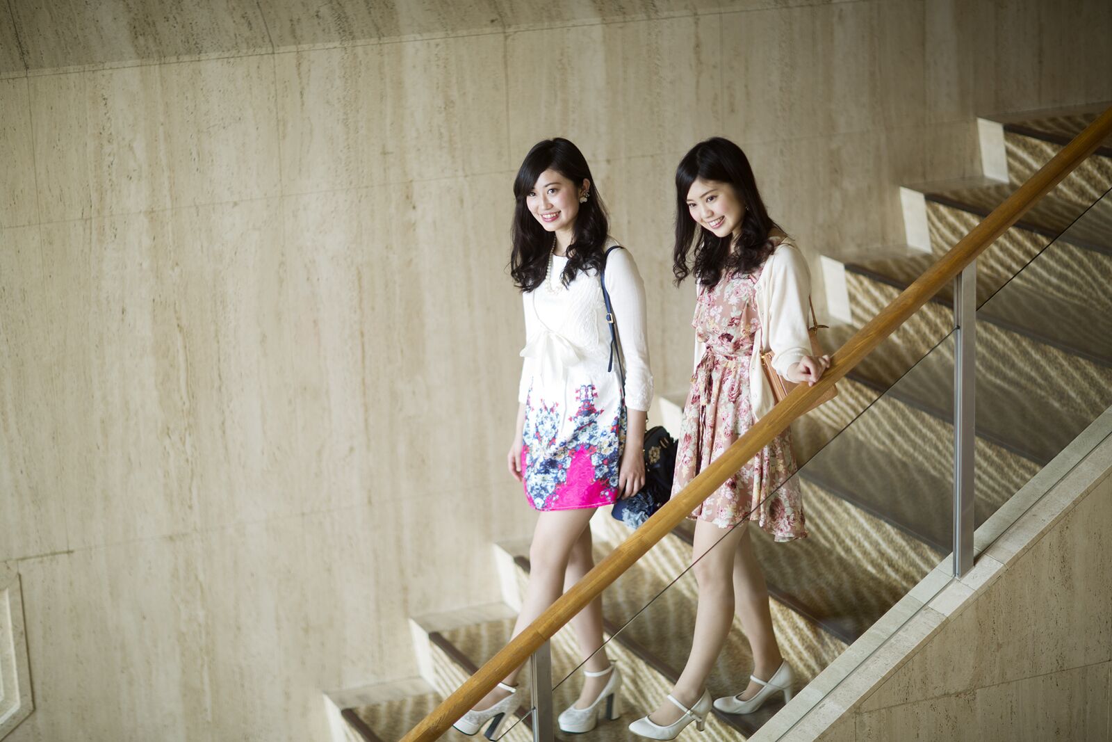 Two individuals descending a staircase with a wooden handrail and a metal railing, set against a plain wall.