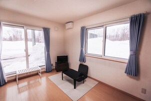 Una habitación con un banco, una silla, un televisor sobre una cómoda y ventanas amplias con cortinas.