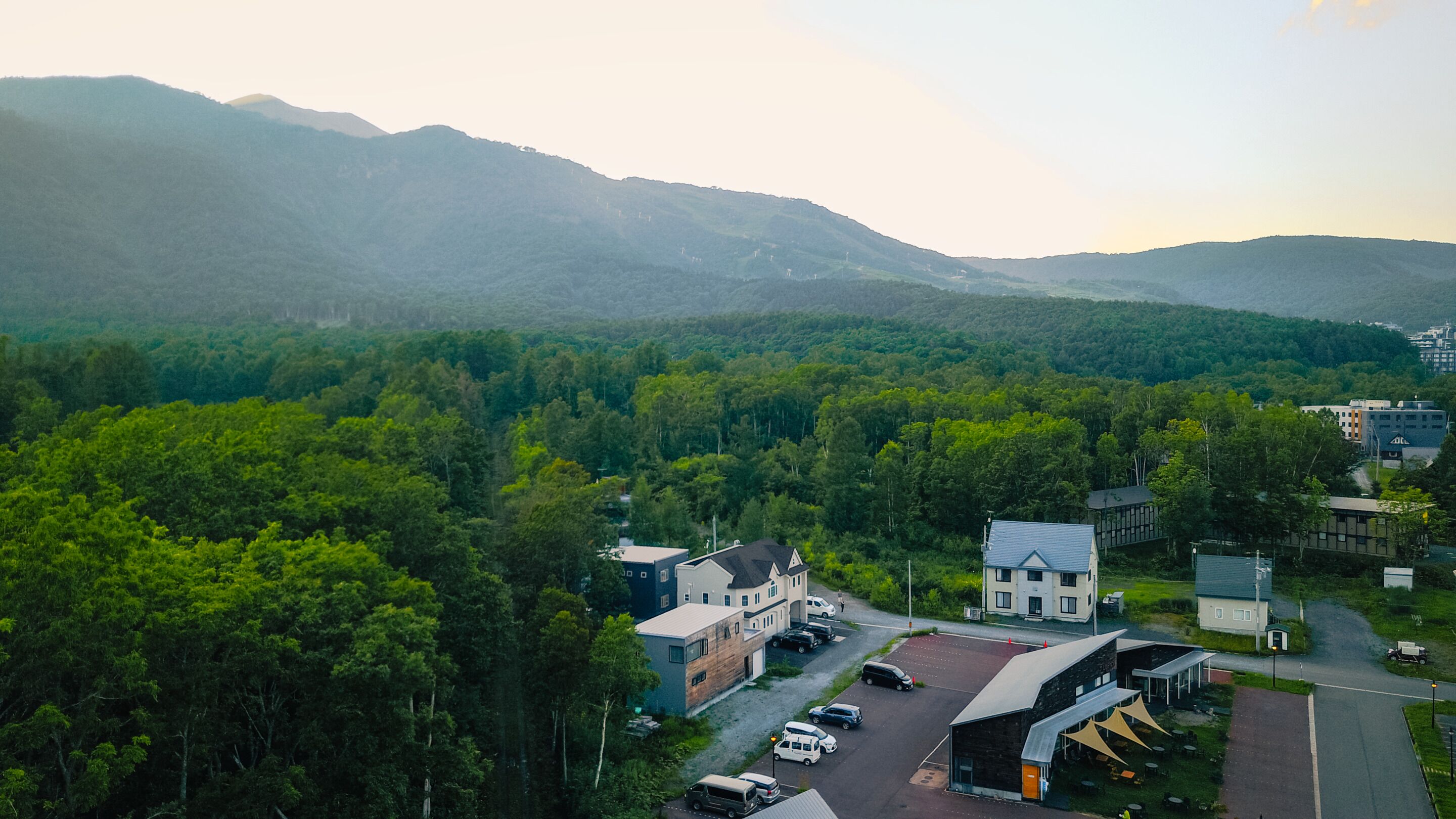 Vista aérea de un pueblo con edificios rodeados de un bosque denso y montañas al fondo.