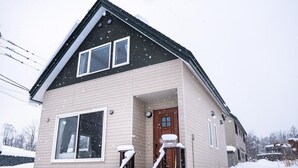 A two-story house with a brown door and windows, surrounded by snow. - Trailside Apartments & Chalet (Kutchan)